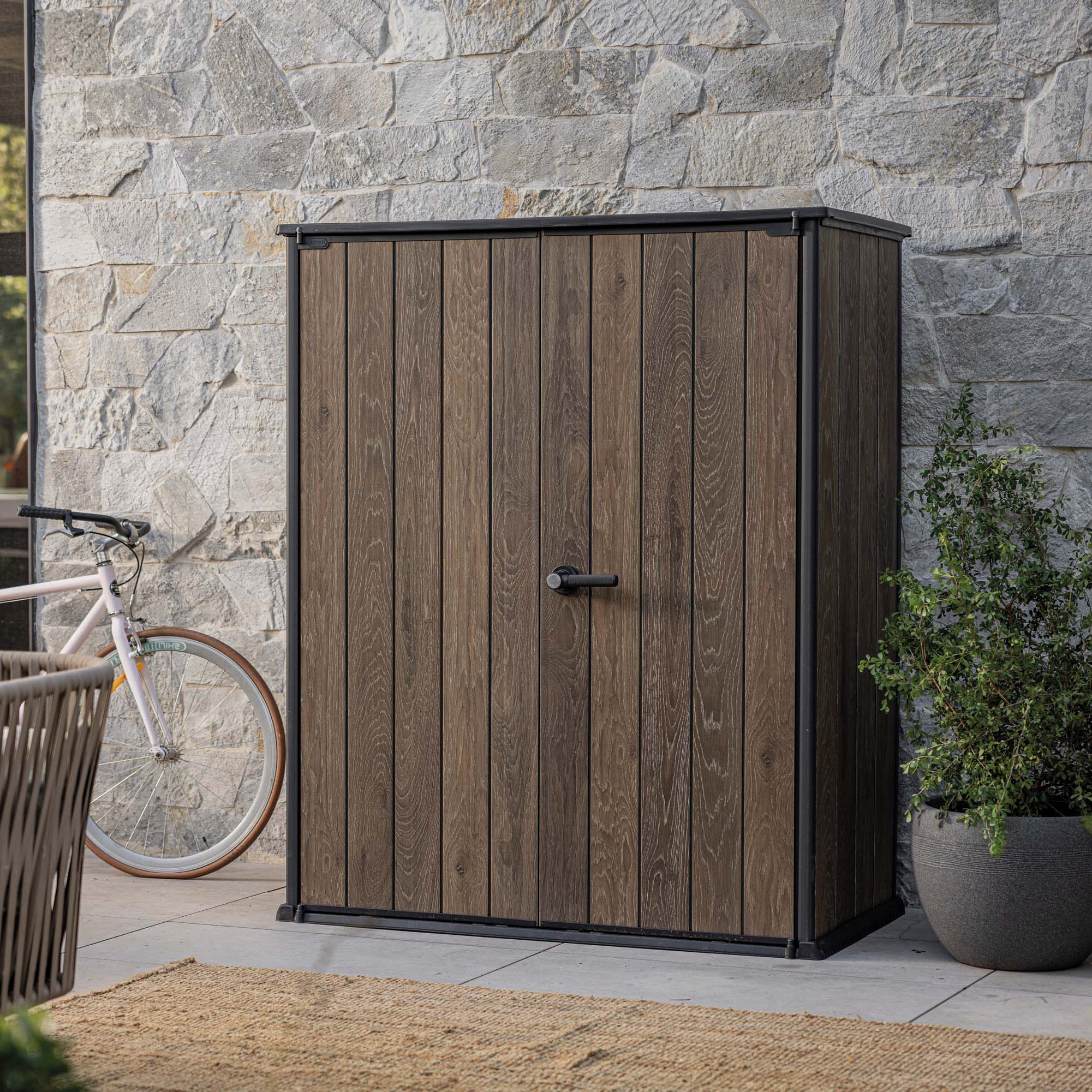 Szafa ogrodowa Deco Vertical 1400L Walnut Keter w drewnianym kolorze ustawiona na tarasie przy kamiennej ścianie