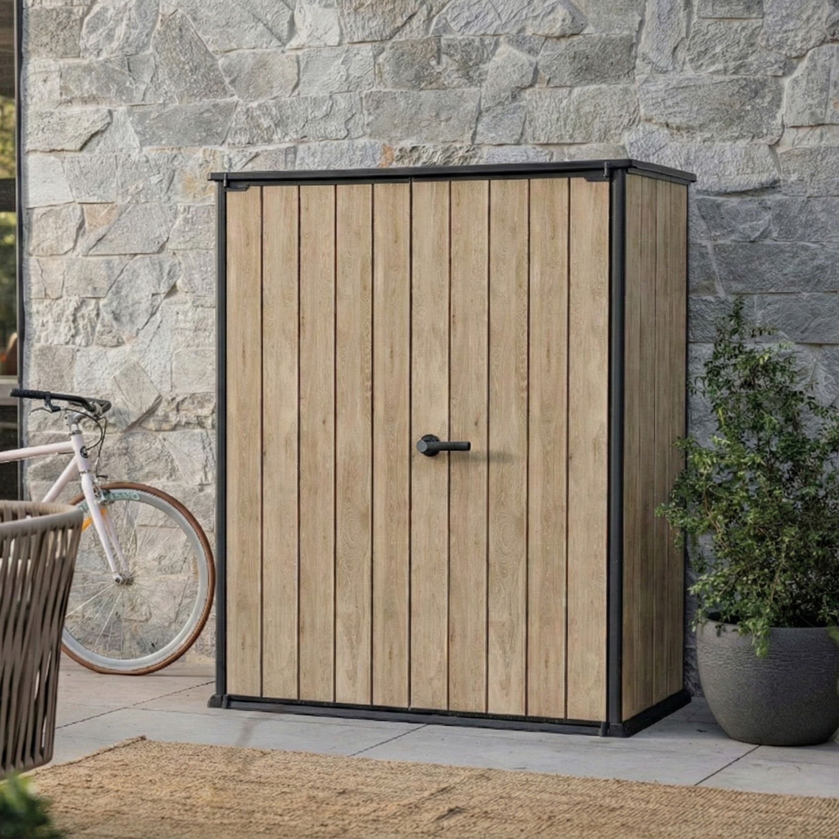 Szafa zewnętrzna Deco Vertical 1400L Ashwood Keter w kolorze jasnego drewna z czarnymi elementami na tle kamiennej ściany