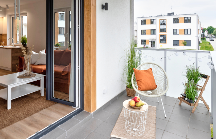 balkon w bloku wylożony szarymi płytkami z mały stolikiem balkonowym i fotelem i kwietniczkiem z ziołami